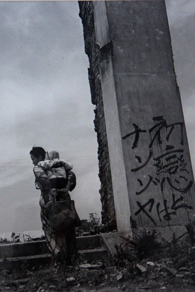 Người phụ nữ địu con đứng trông ngóng bên cạnh một đống hoang tàn. Đất nước Nhật Bản đã gần như bị tàn phá hoàn toànởi cả thập kỷ chiến tranh và hằn sâu vết sẹo của trận đánh bom nguyên tử ở Hiroshima và Nagasaki.